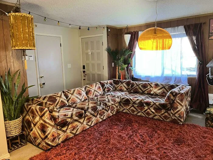 A cozy living room featuring a large patterned sectional sofa, characteristic of 1970s and 1980s interior design.