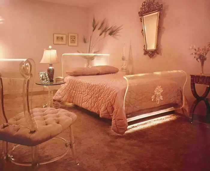 A stylish 1970s bedroom interior design with a pink bed, mirrored headboard, and clear acrylic furniture.