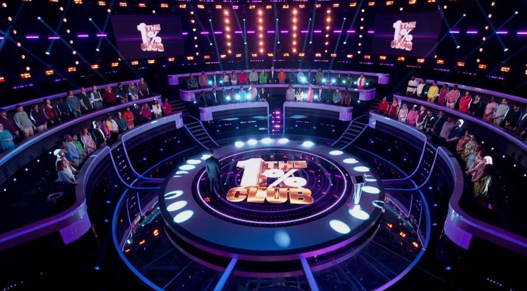 A game show set for The 1% Club, with a host standing on a circular stage and contestants seated in tiers.