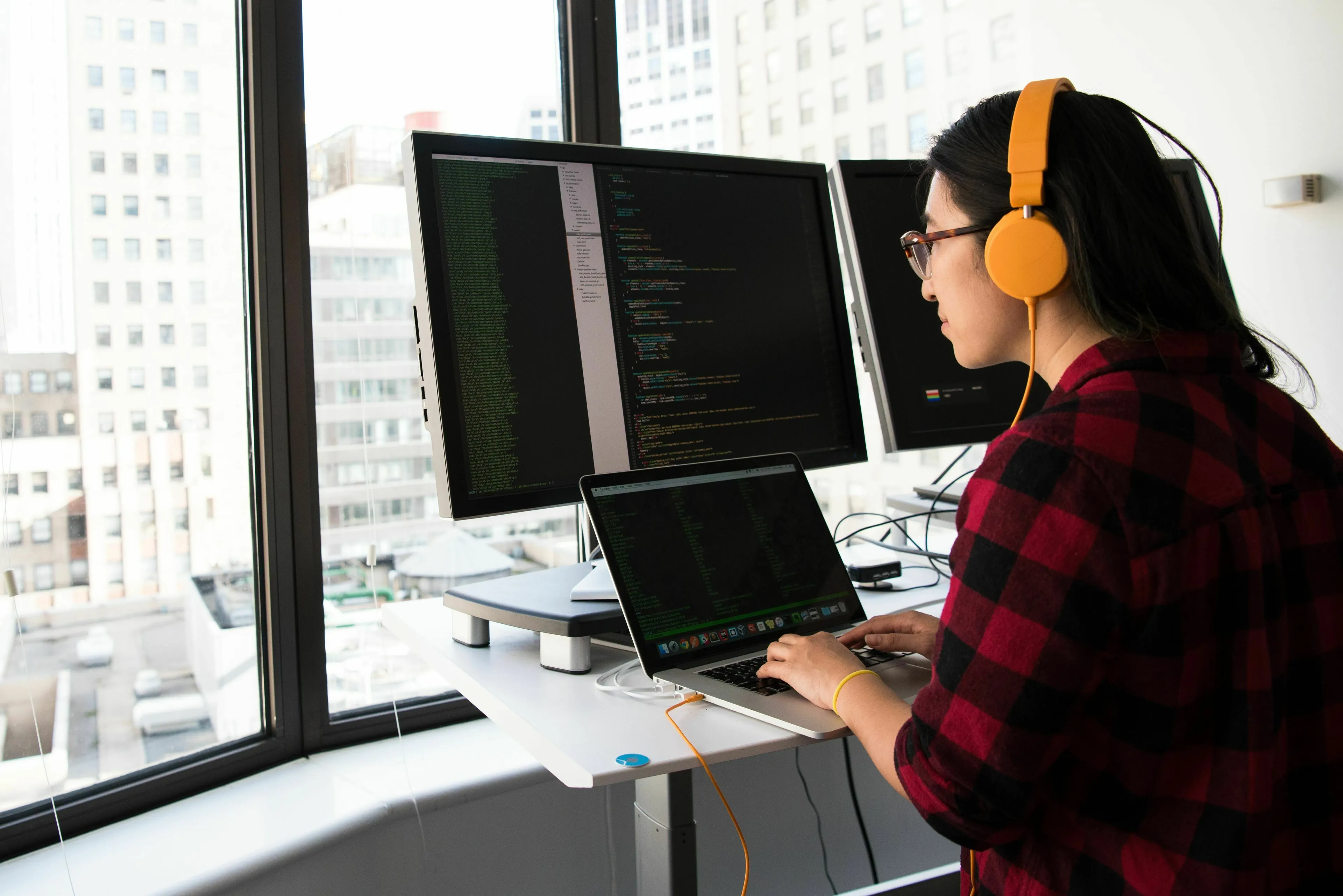 A woman wearing headphones and glasses, looking at multiple computer monitors with code, choosing the odd one out.