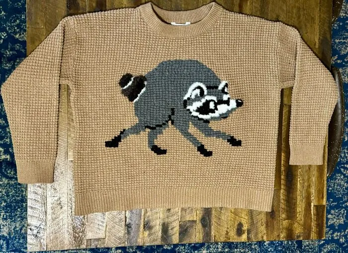 Beige crochet sweater with raccoon design laid flat on wooden surface