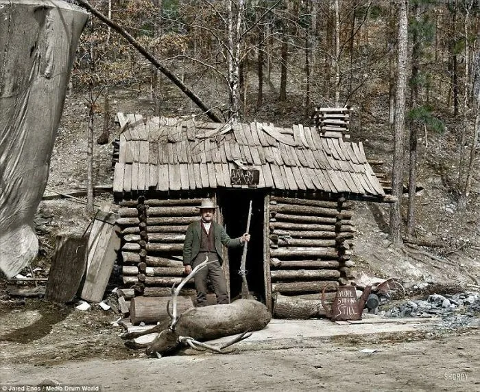 Historical photos: A man standing proudly with his hunting rifle and a killed deer outside a rustic log cabin in the woods.