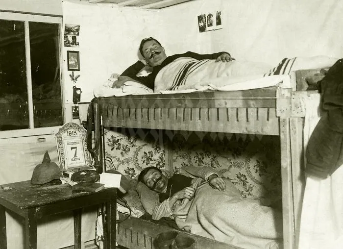 A historical photo of two men with moustaches resting in bunk beds, with a calendar showing March 7, 1915.