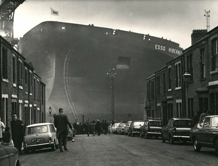 Historical photo of a huge ship, Esso Hibernia, dwarfing a street with parked cars and onlookers, a vivid past scene.