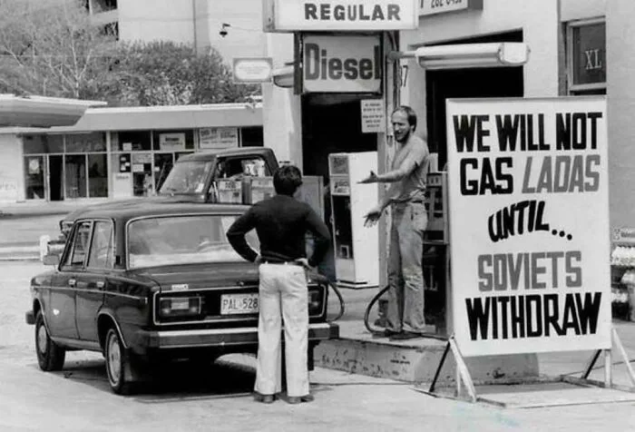Historical photo of a man refusing to gas a Lada car with a sign that reads, WE WILL NOT GAS LADAs UNTIL SOVIETS WITHDRAW, a moment from the past.