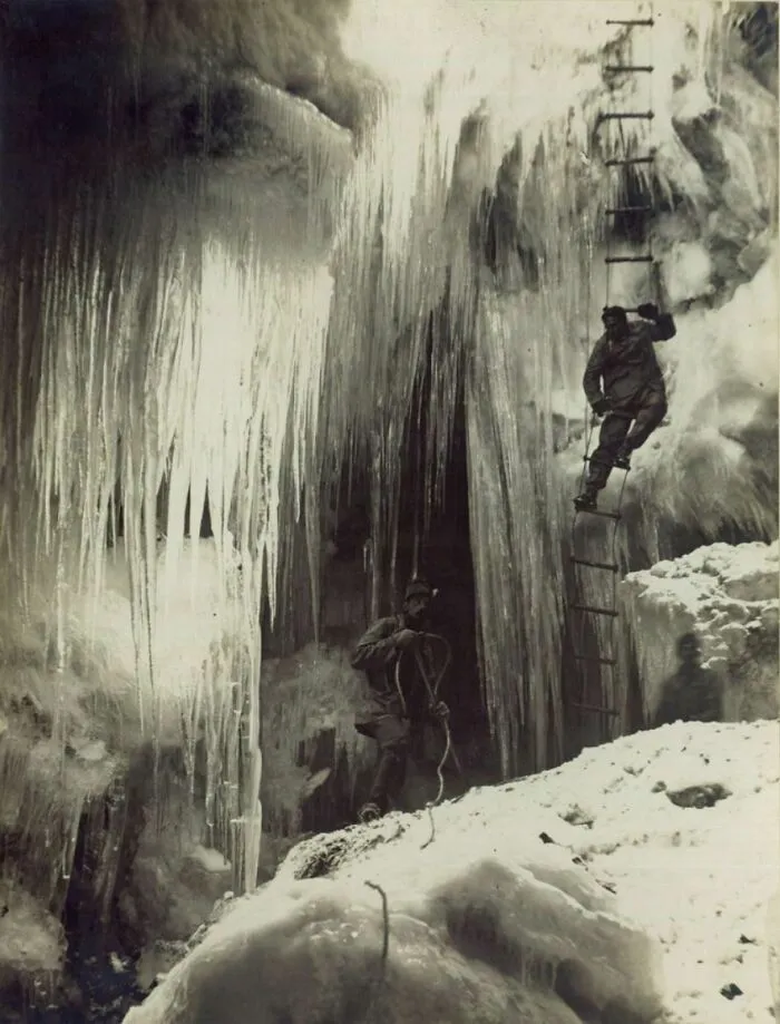 A historical photo of two climbers on an icy mountain, one ascending a ladder, documenting a challenging past expedition.