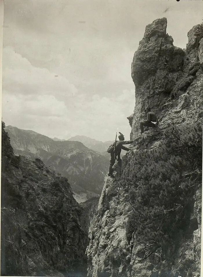 A striking historical photo captures two soldiers scaling a steep, rocky mountain face amidst a vast, rugged landscape.