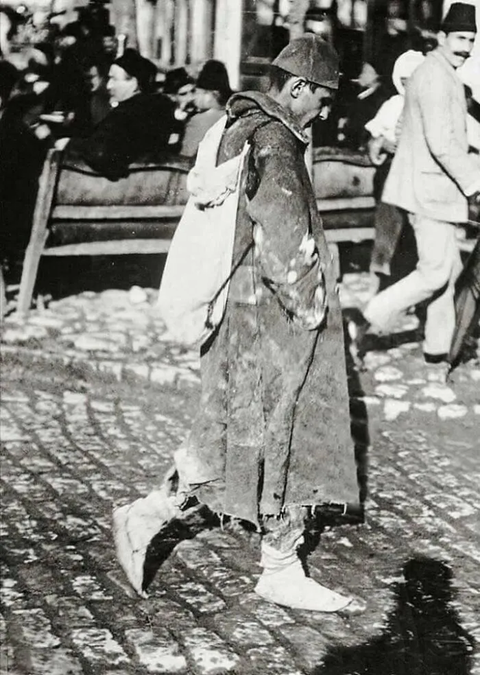 A man in worn clothing and a cap walks down a cobblestone street, carrying a sack, in a historical photo of a past era.