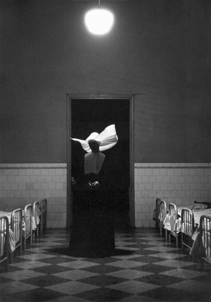 A nun in a habit stands in a dimly lit hallway between rows of beds in a historical photo of a past hospital.