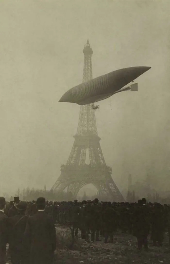 A historical photo of a dirigible flying above the Eiffel Tower with a crowd below, remembering a past event.