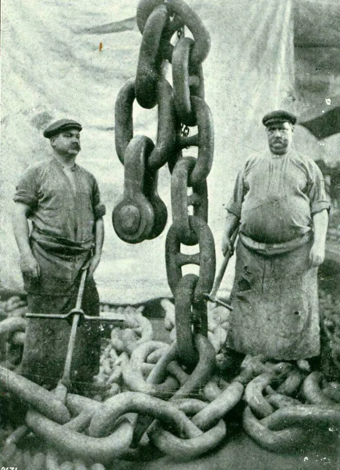 Two strong men standing beside a colossal anchor chain, highlighting a fascinating historical photo.