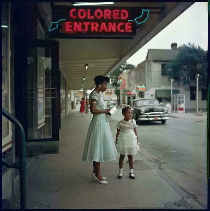 Historical photo showing a woman and a young girl standing on a street, with a neon sign for a segregated entrance.