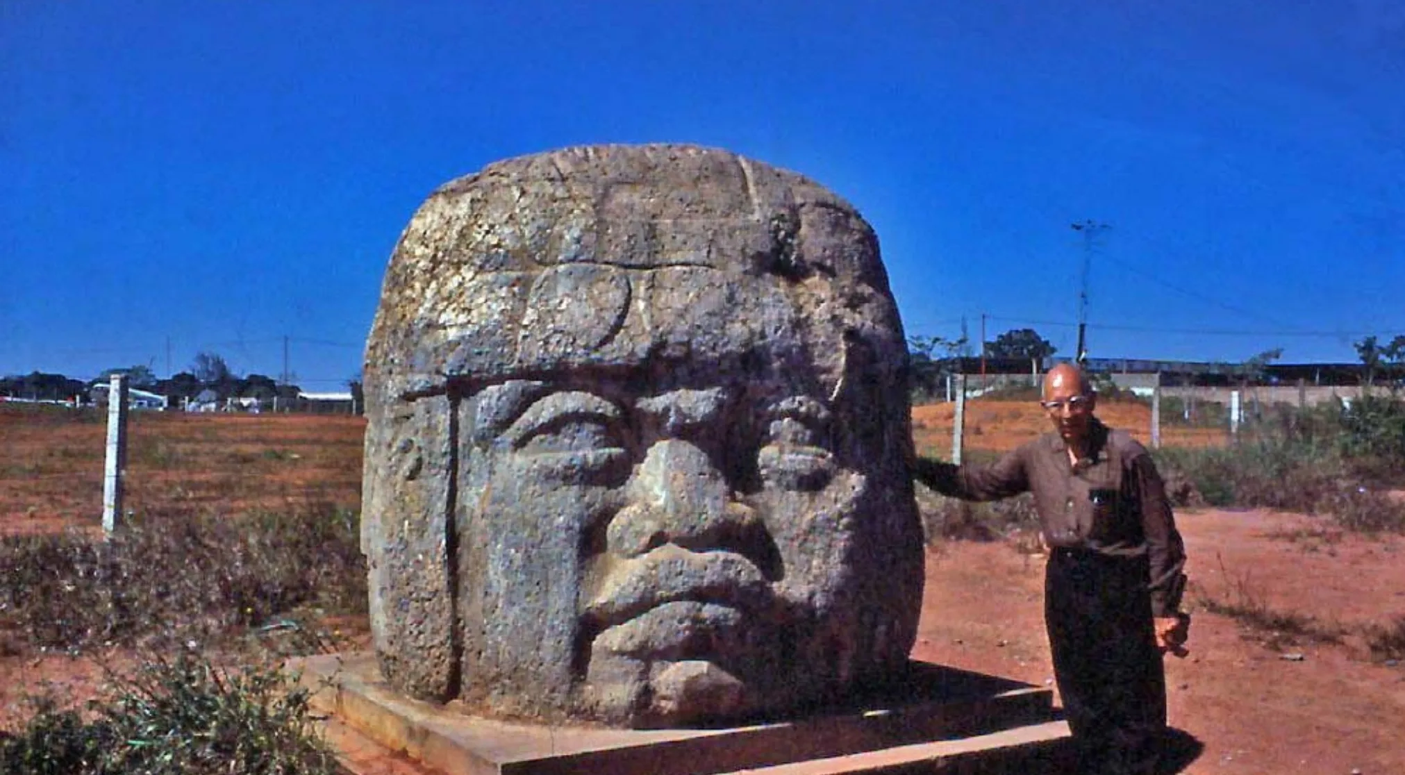 Man next to a colossal stone head from an ancient civilization, under a bright blue sky.