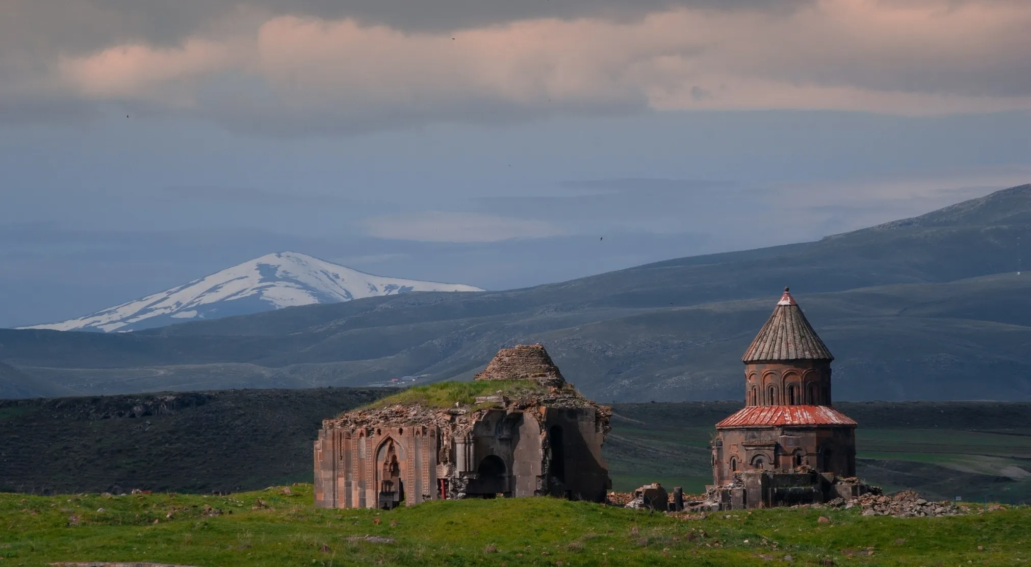 Ancient city ruins of two historic churches in a green landscape with mountains and a snowy peak under cloudy skies.