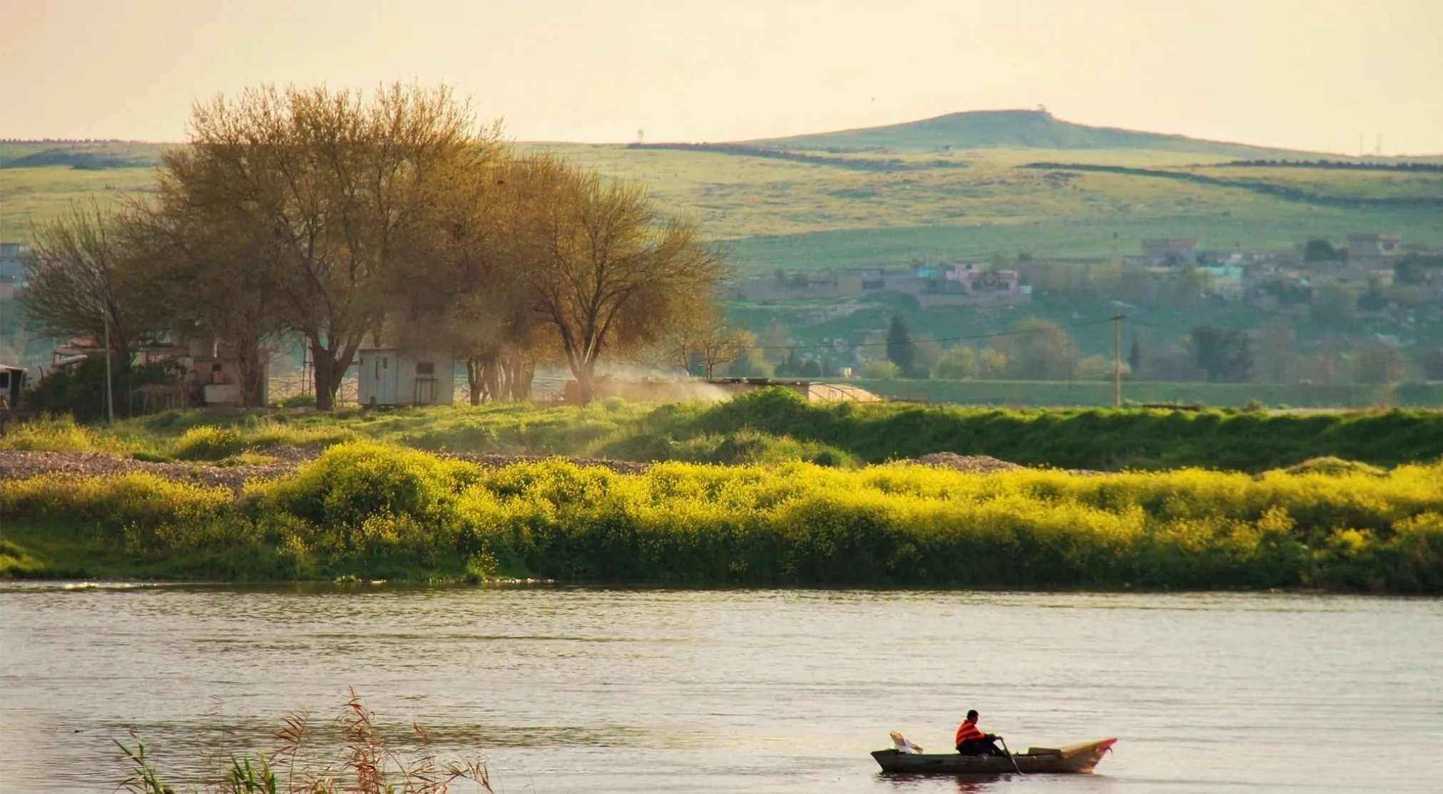 Person rowing a small boat on a river, with green hills, scattered houses, and bright yellow flowers. Ancient Mesopotamia rivers.