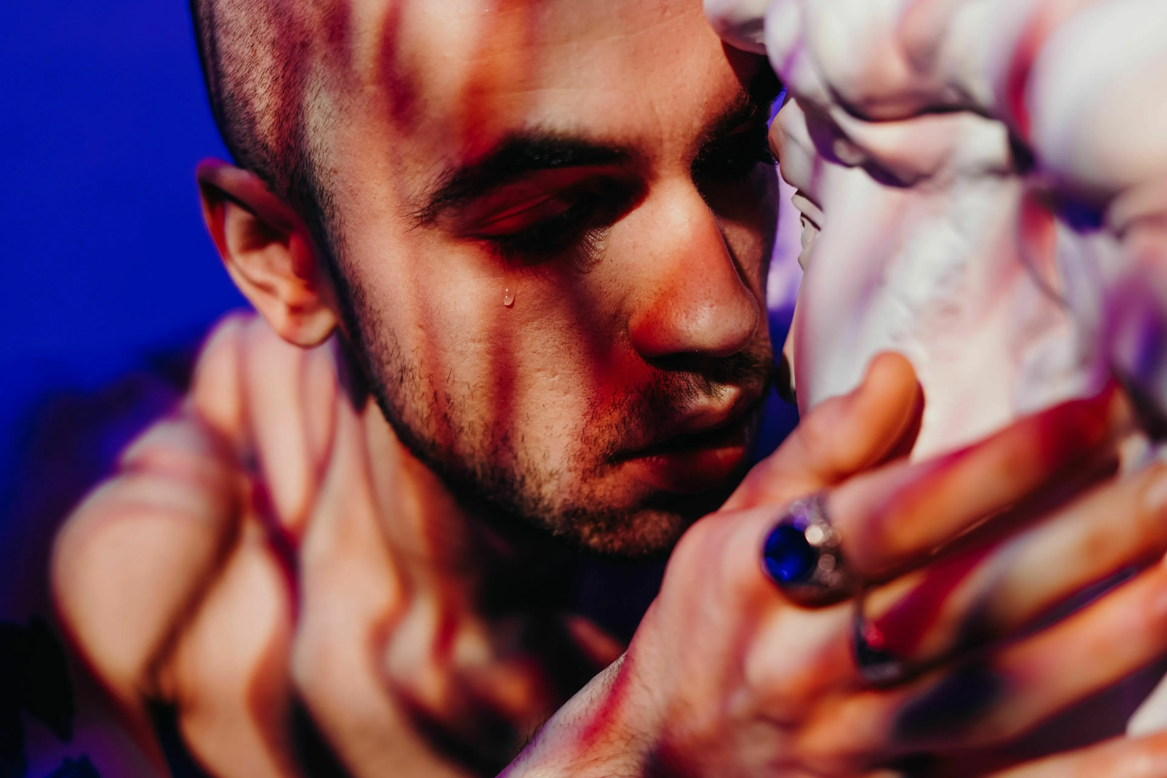 A man with a tear on his cheek, lit by red and blue light, holding a white statue. A sympathetic villain.