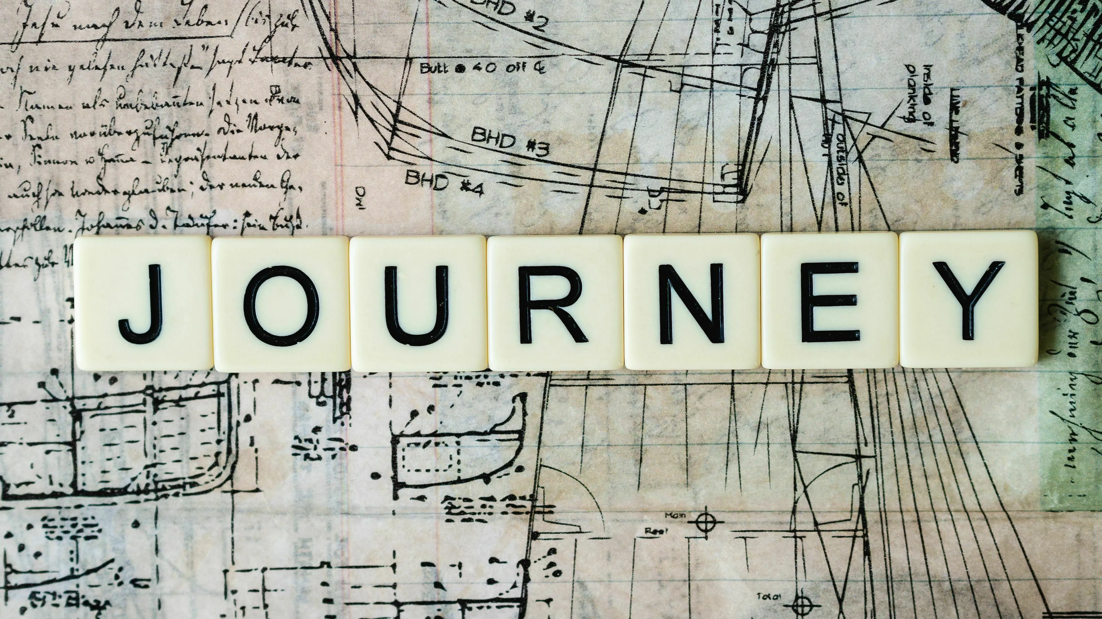 Scrabble tiles spell JOURNEY over mythological quest blueprints, representing a destination.