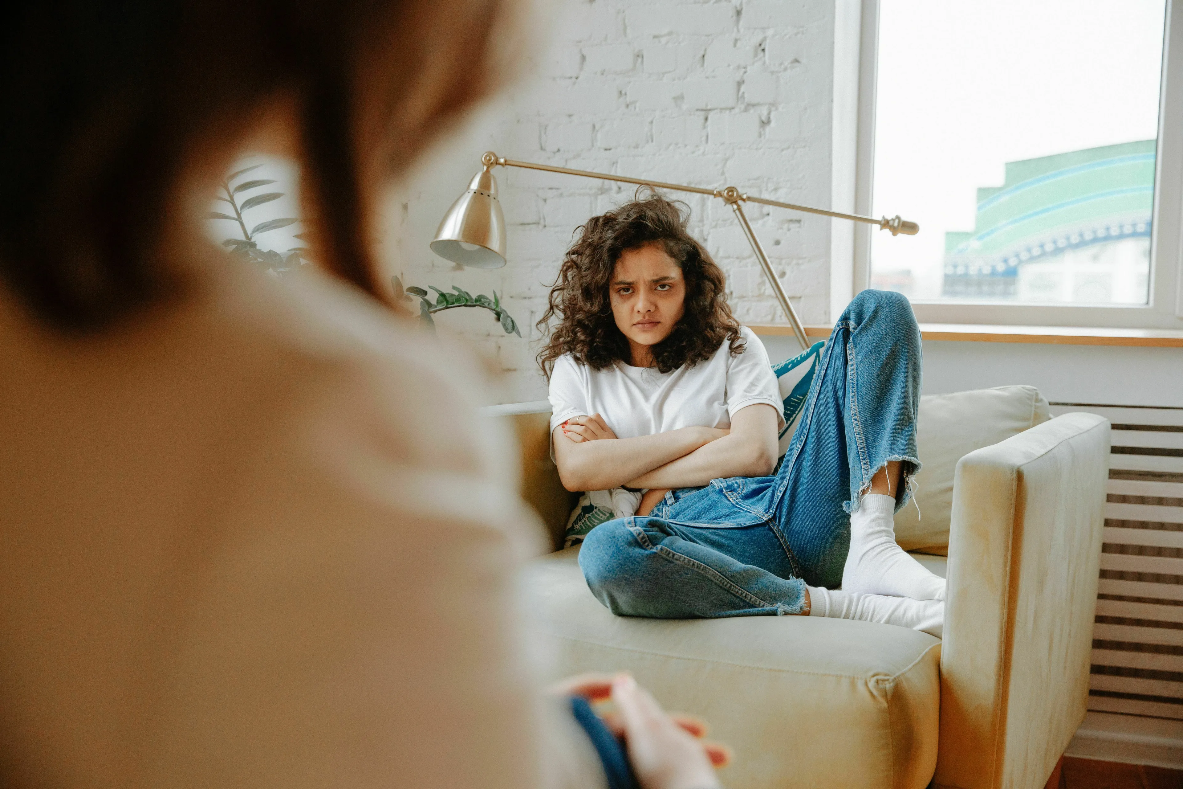A woman with curly hair sits on a couch, arms crossed, looking at someone off-camera, representing a green flag for catching a friend in a small lie.