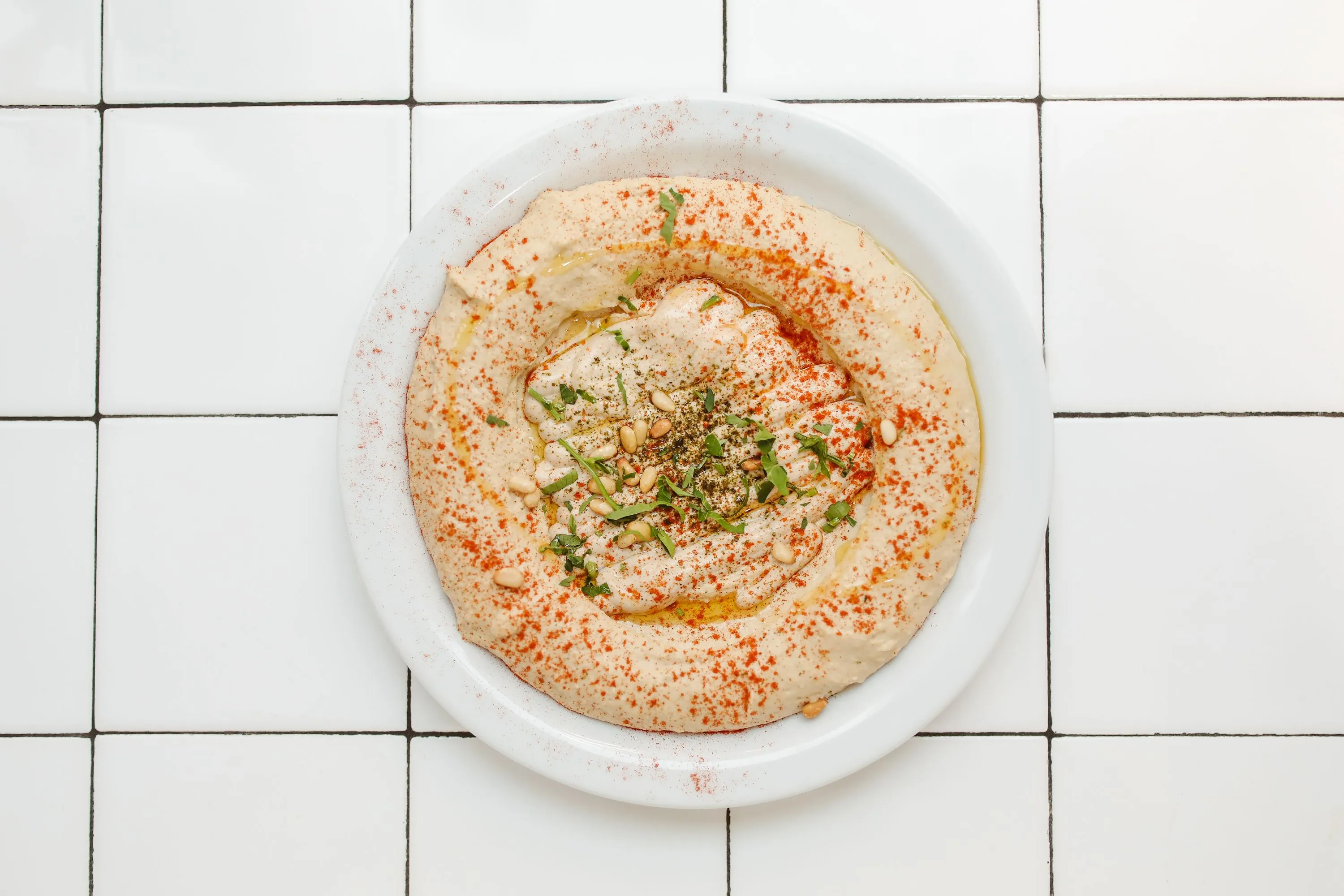 A dish of homemade hummus, garnished with paprika, parsley, and pine nuts, sits on a white tiled surface.