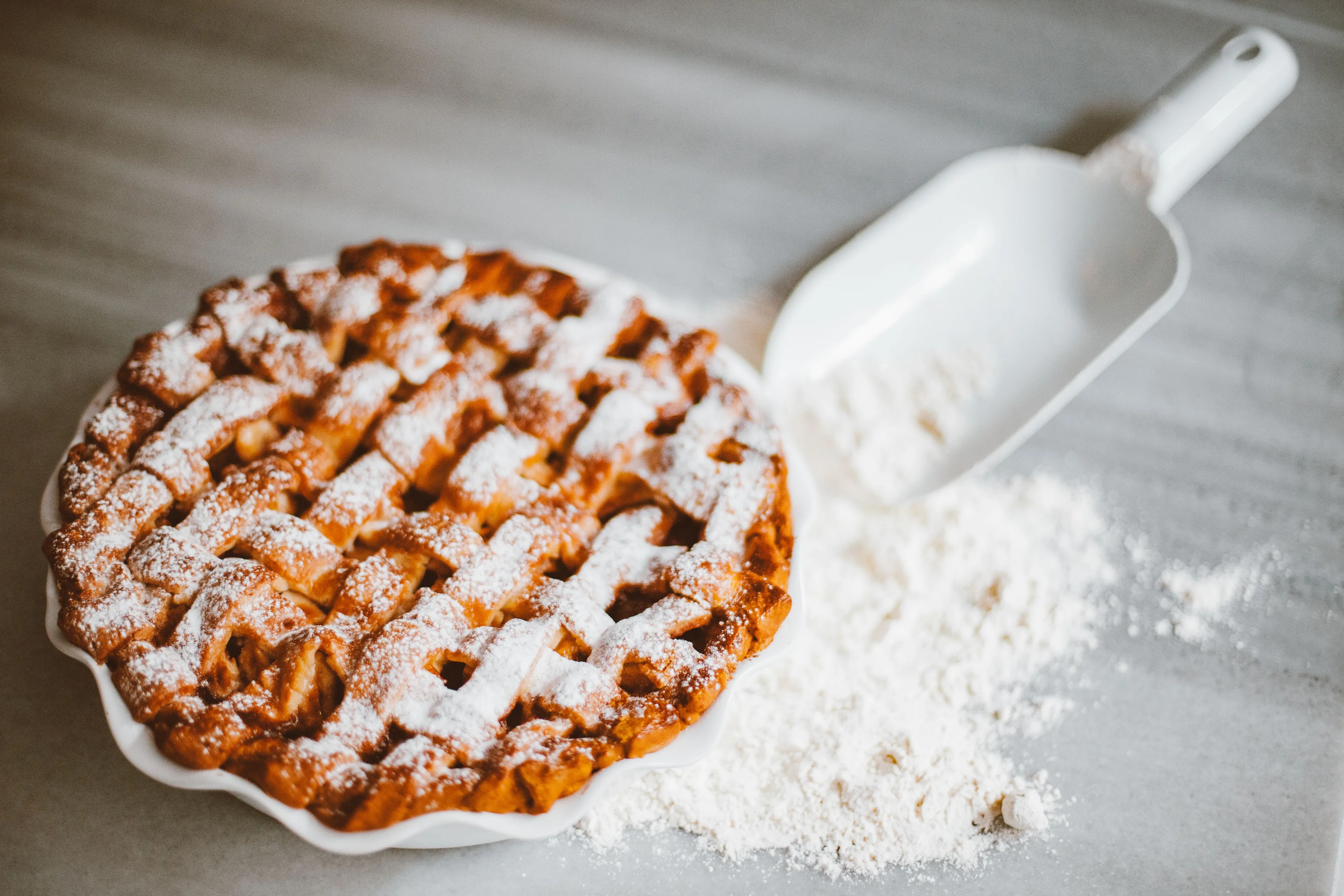 A freshly baked apple pie with a lattice crust, dusted with powdered sugar, next to a pile of flour.