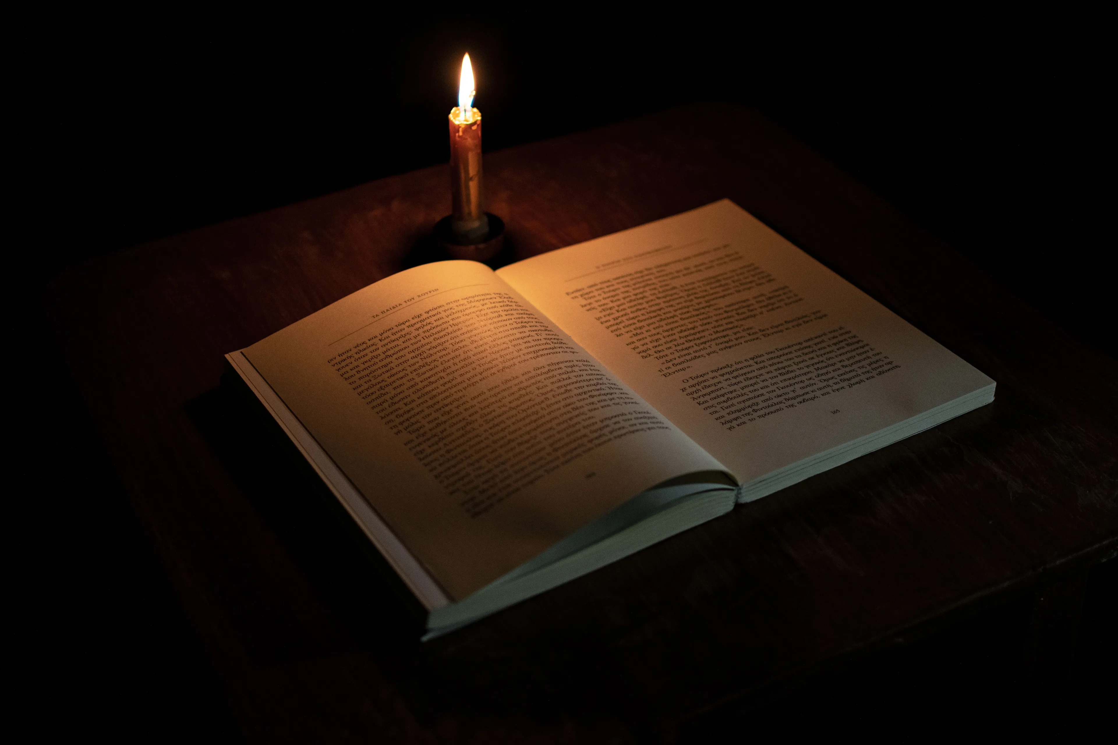 Last thing you do before bed: An open book illuminated by a single lit candle on a dark wooden table, for reading.