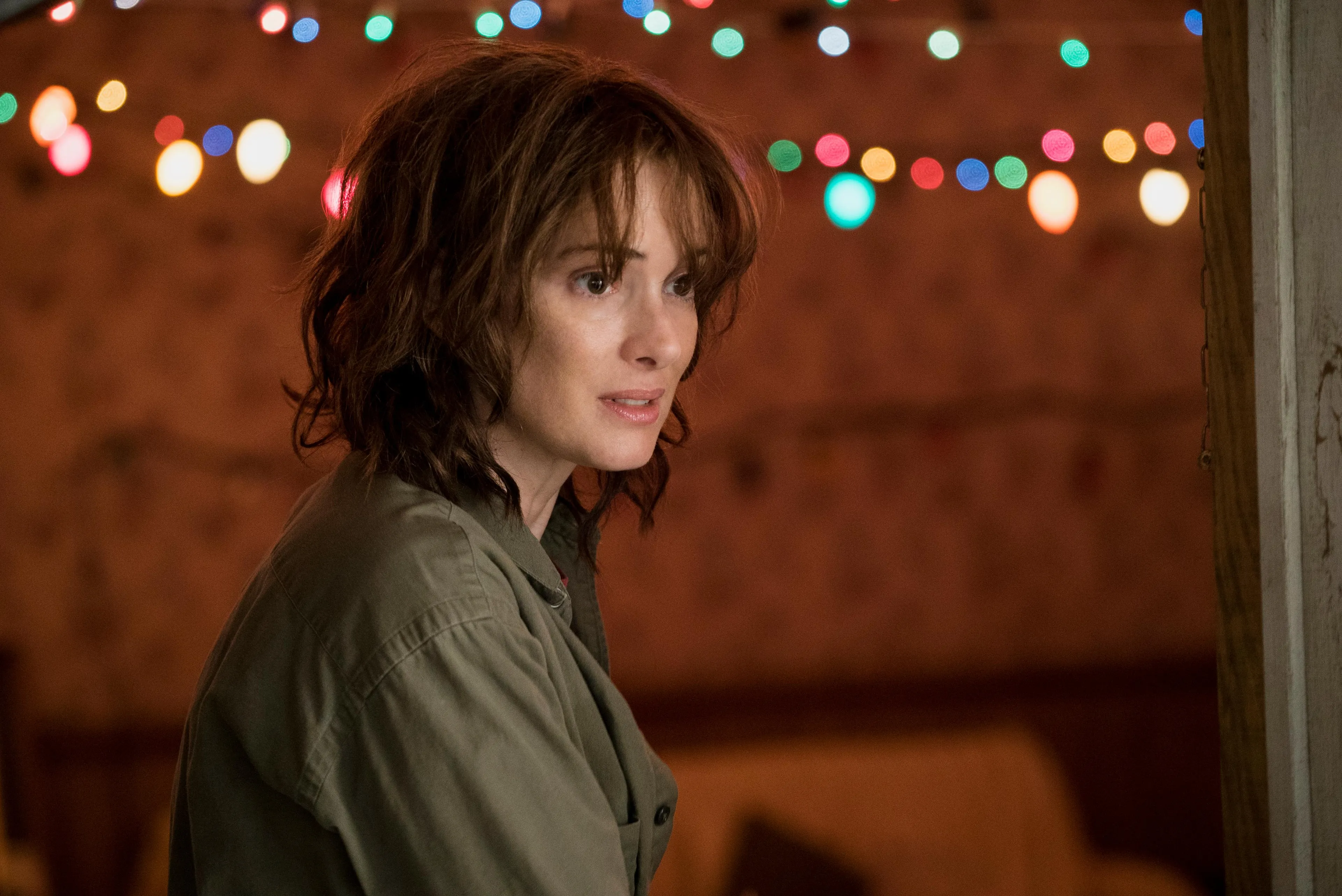 Joyce Byers, a fictional mentor, looking pensive in front of a wall adorned with colorful Christmas lights.