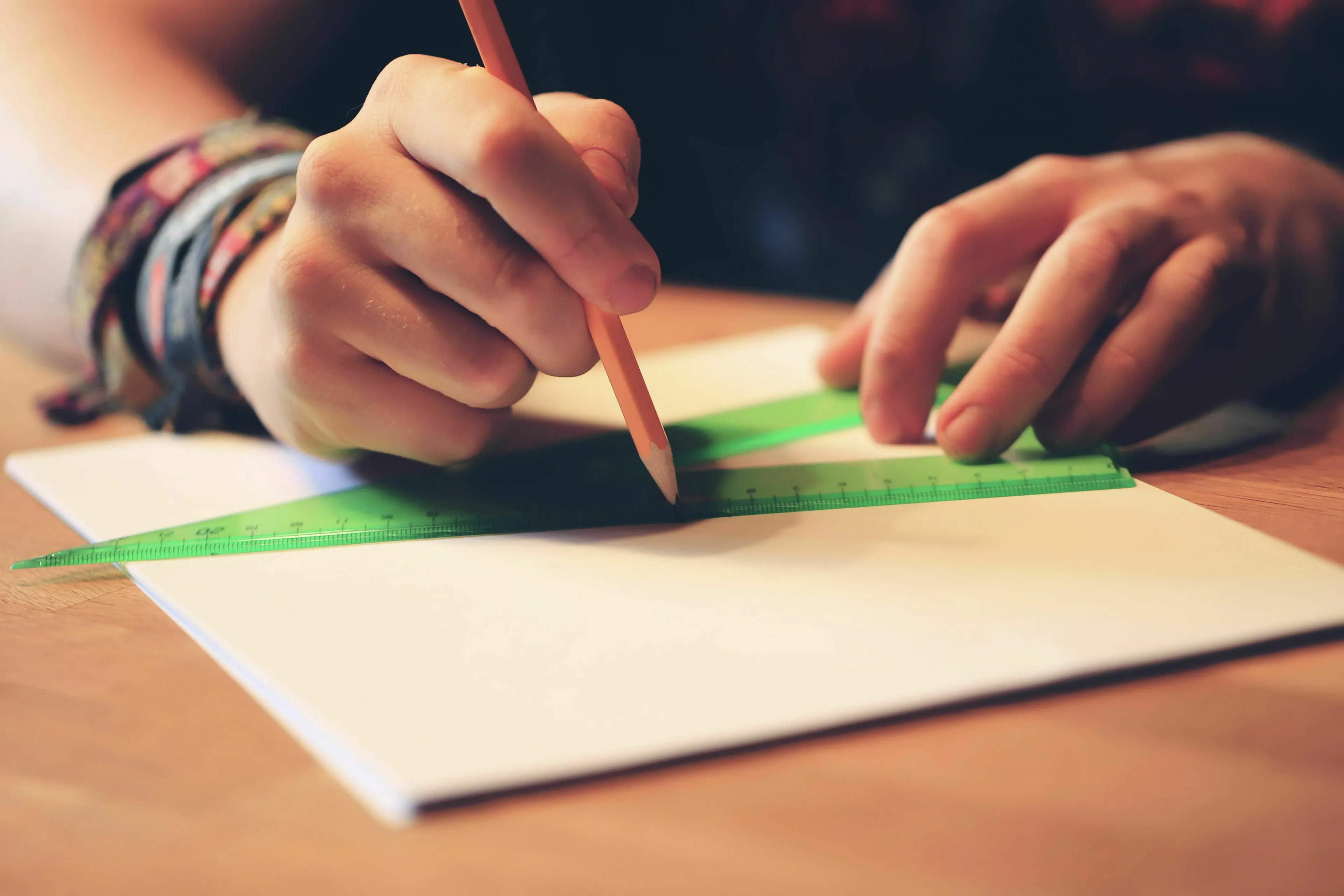 Hands drawing a straight line with a pencil and green ruler on white paper, solving how many sides a triangle has.