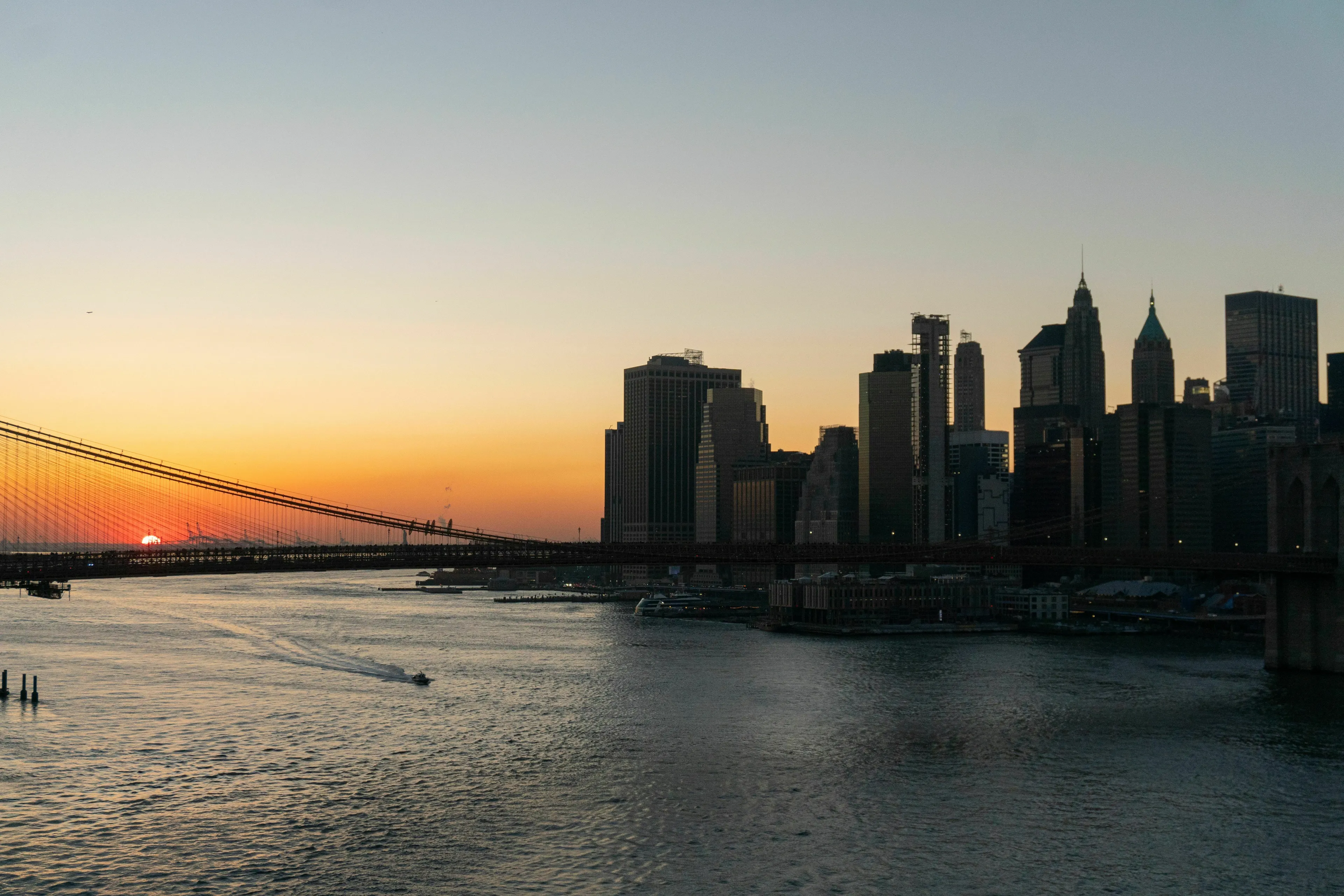 Sunset over New York City skyline and a bridge with a boat on the water.