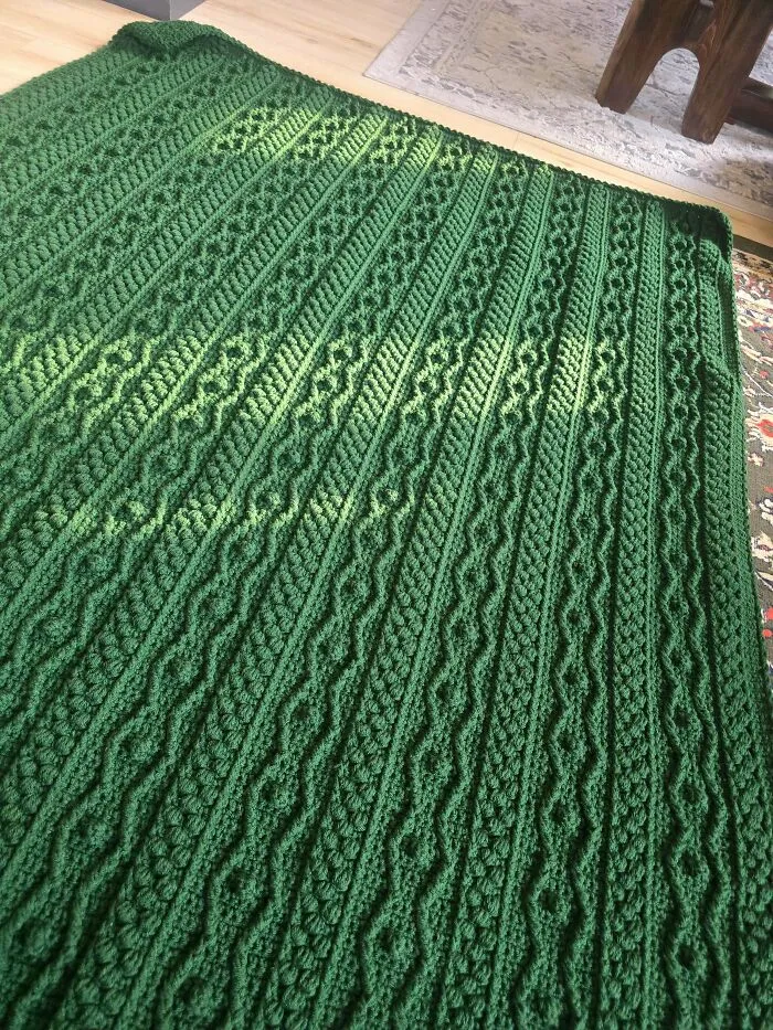 Textured green crochet blanket with cable and diamond patterns on wooden floor