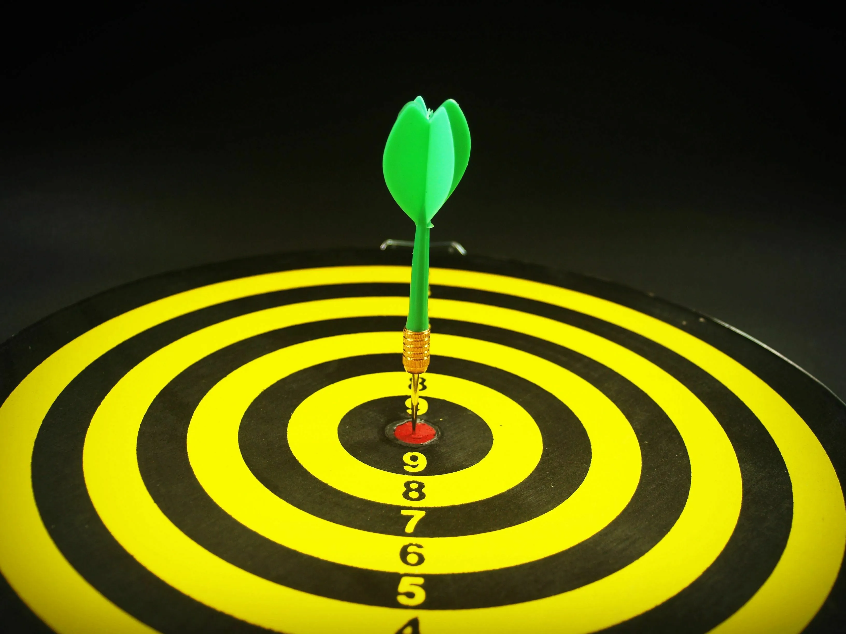 Watching someone else succeed, a green dart hits the bullseye of a yellow and black dartboard, signifying achievement.