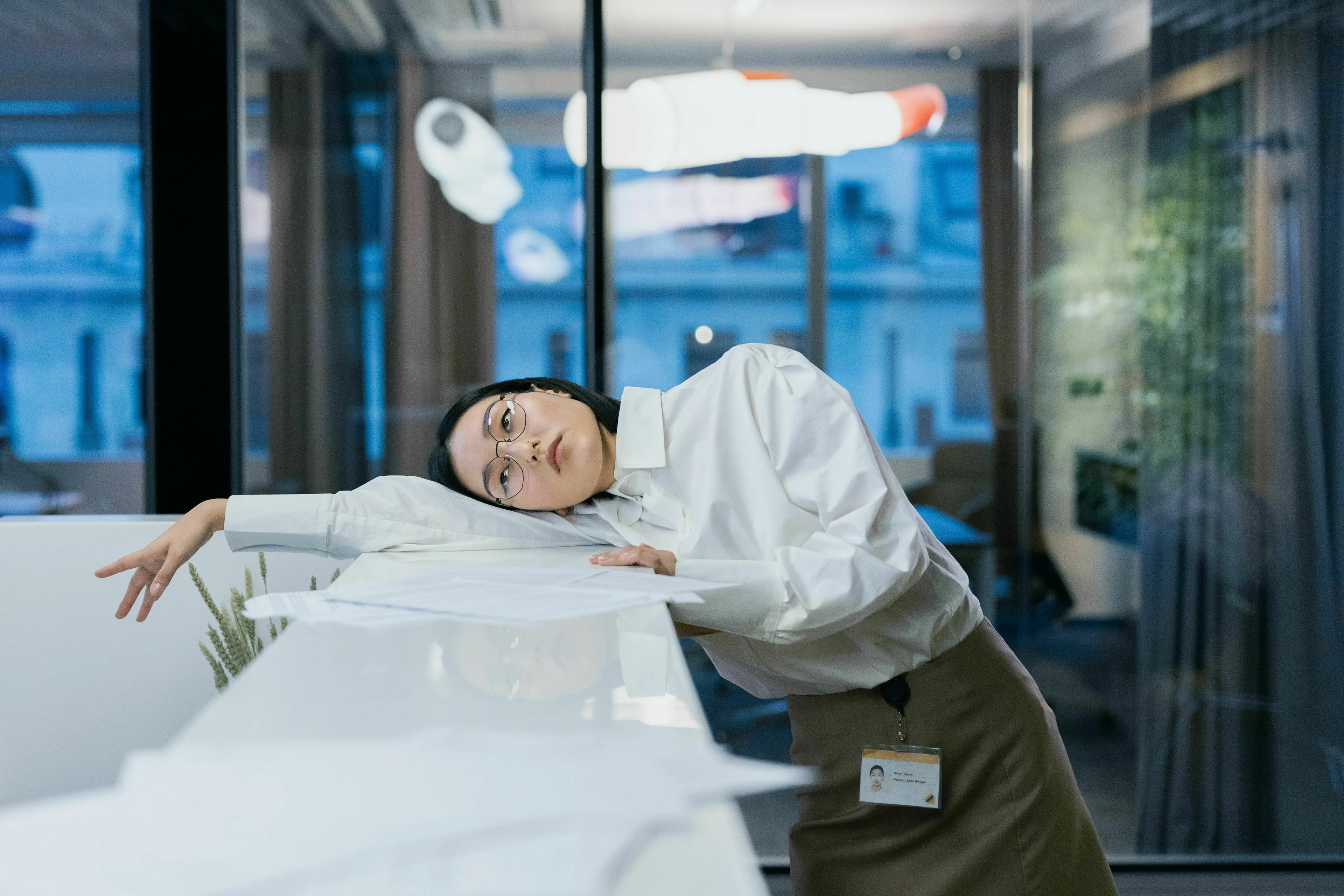 A woman with glasses looks bored at work, leaning on a desk with papers. Her expression depicts what triggers your envy.