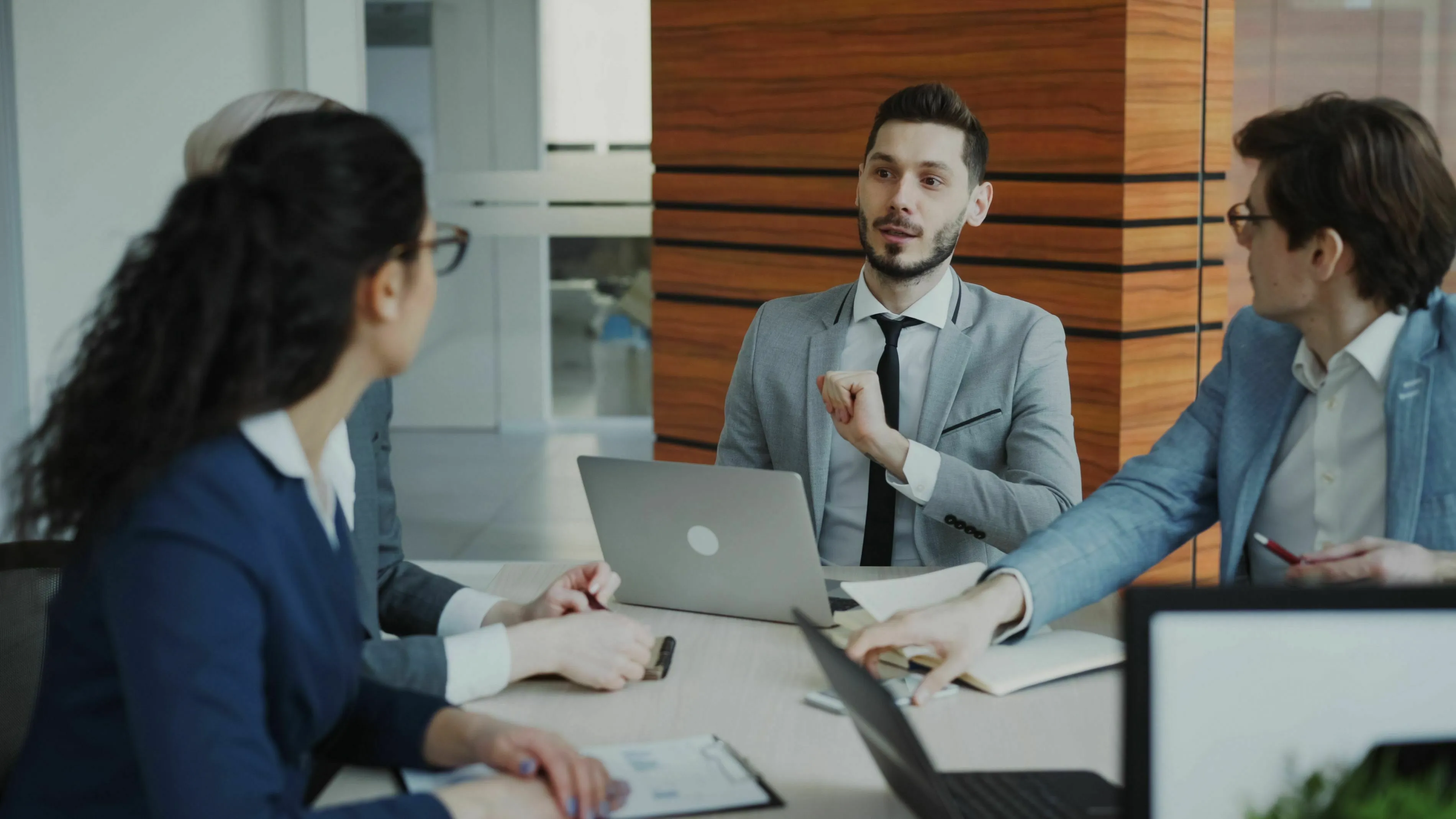 A man in a gray suit speaks in a meeting, surrounded by coworkers. Triggers envy when a coworker gets praised.