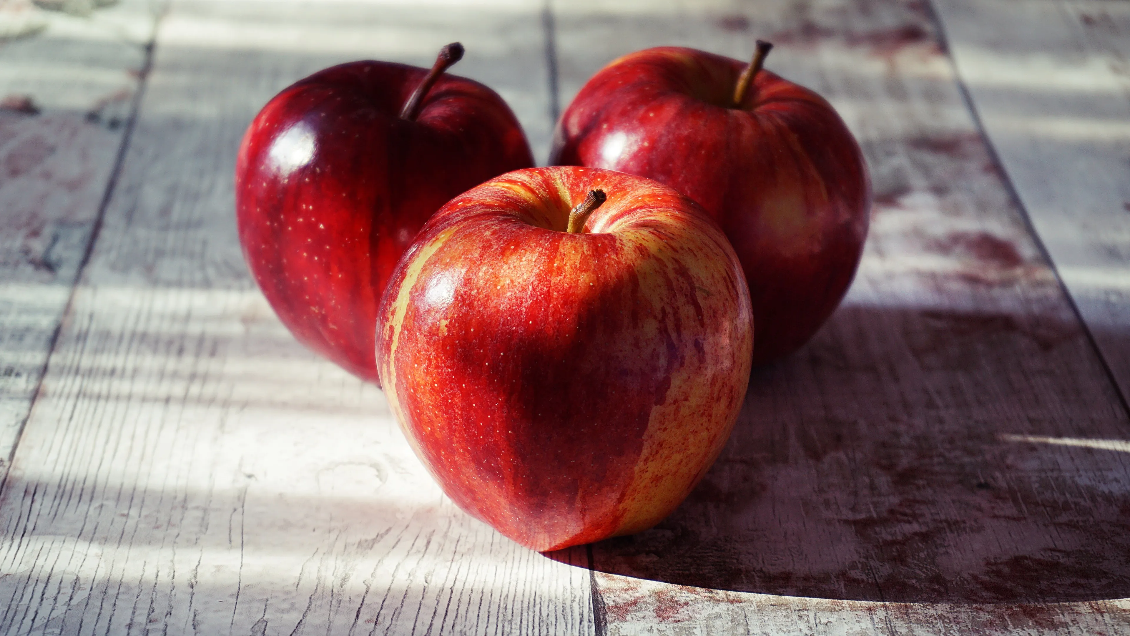 Three red apples with some yellow patches, sitting on a light wooden surface in sunlight, highlighting the choice of apple.