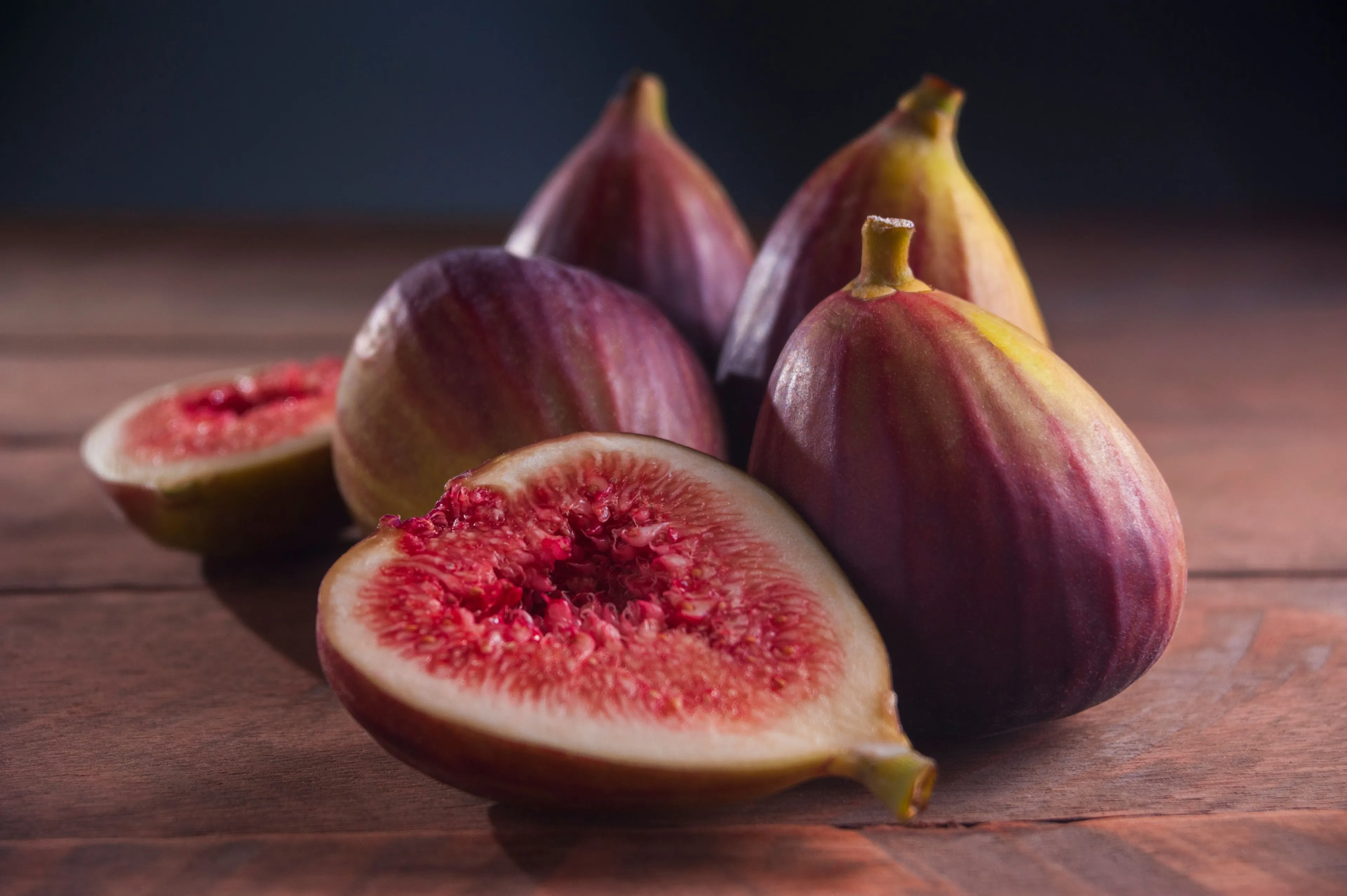A close-up of fresh, ripe figs, some whole and one cut in half, revealing its red interior on a wooden surface.