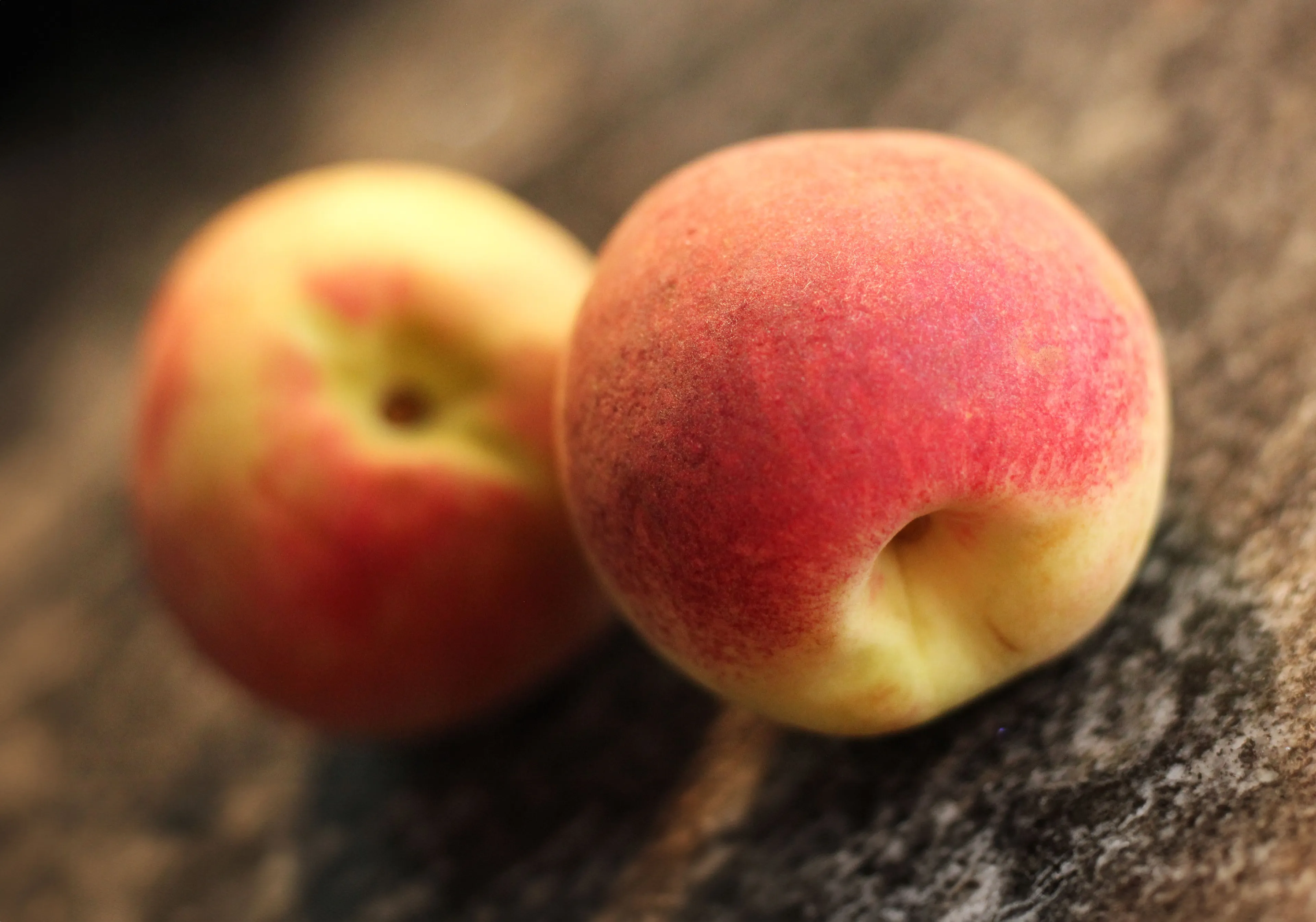 Two ripe peaches, one in focus showing its soft, fuzzy skin with red and yellow hues, on a stone surface.