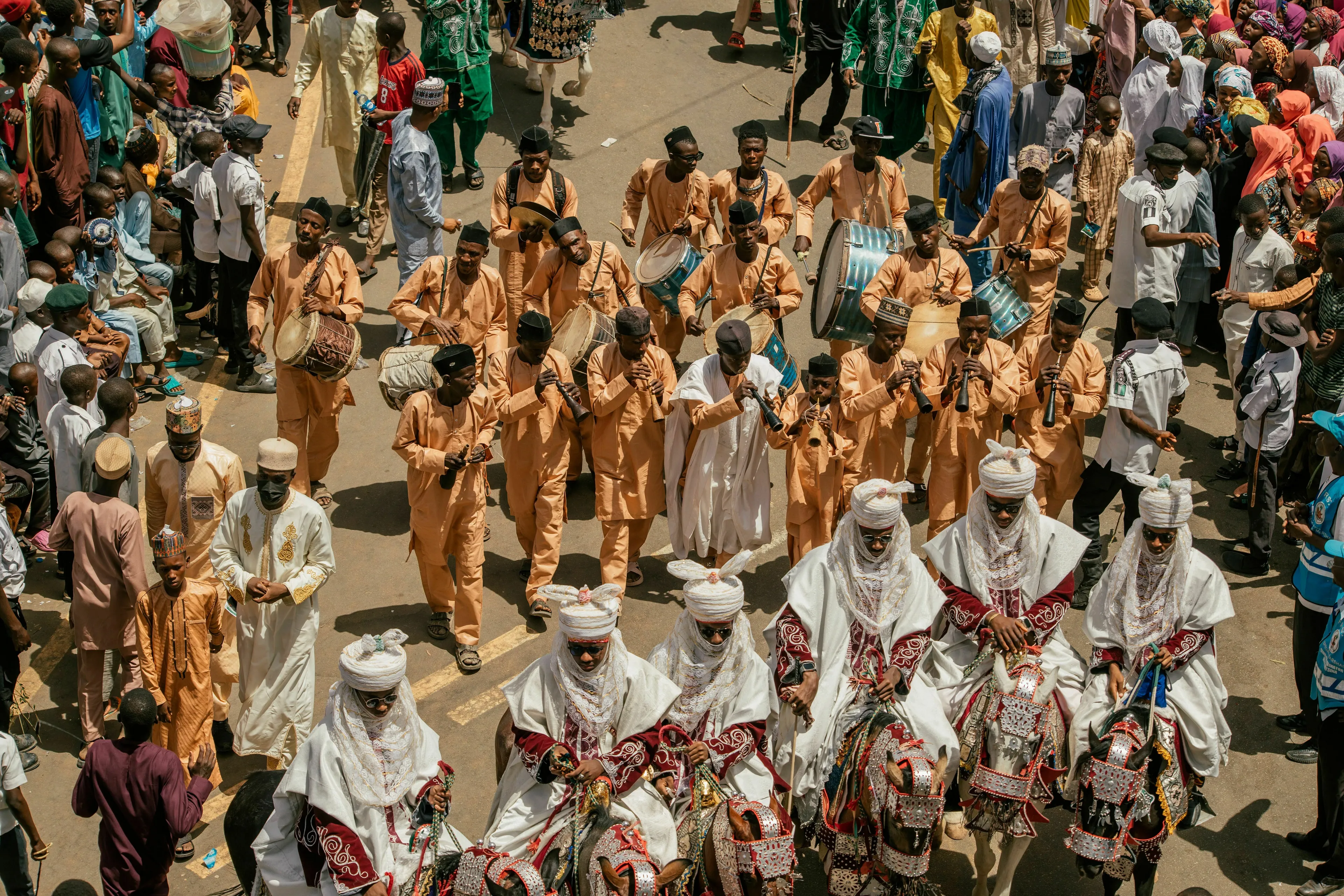 A vibrant traditional parade with people in orange garments playing instruments and men on horseback, showcasing tradition.