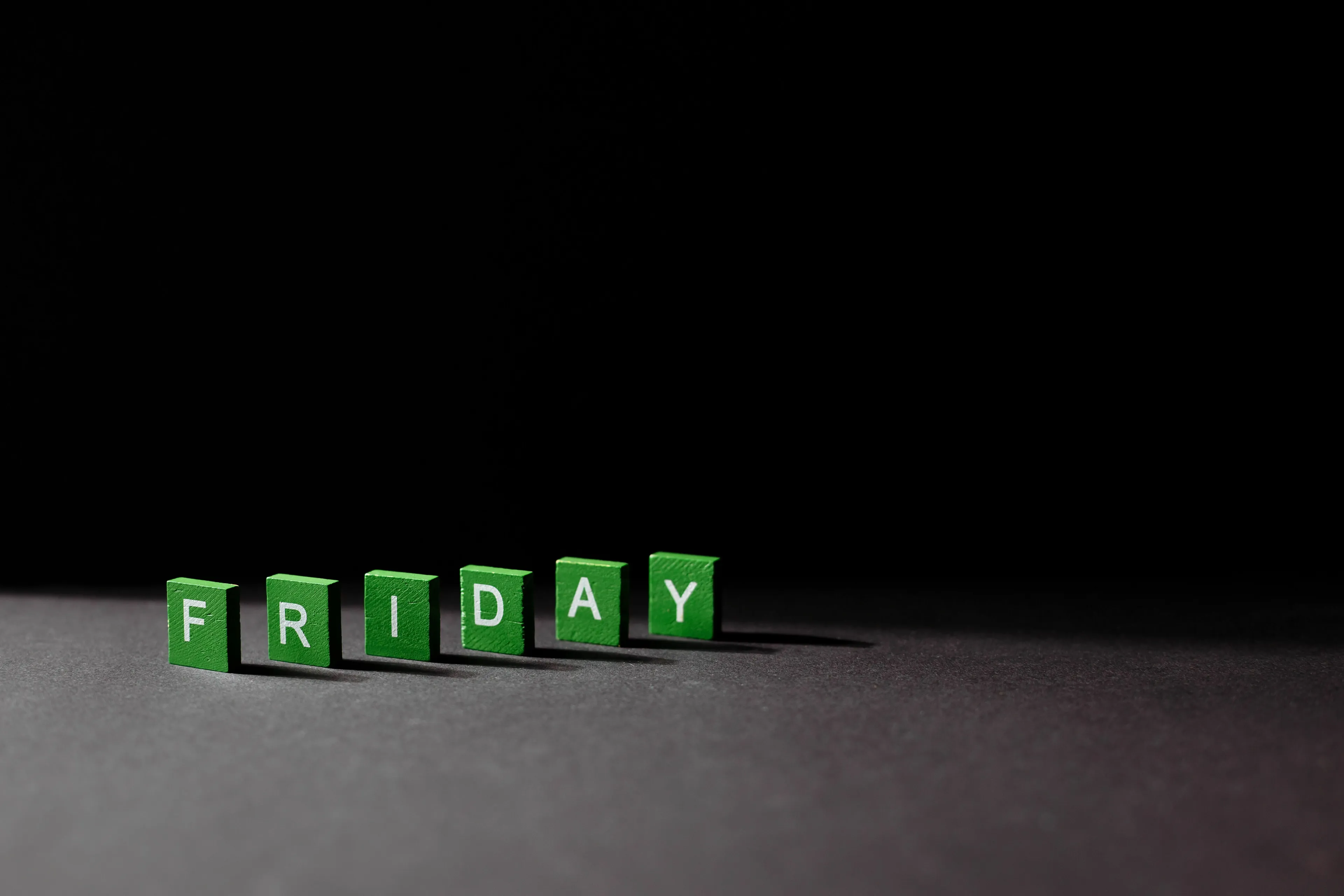 Green letter tiles spelling FRIDAY are arranged on a dark surface, symbolizing the ideal Friday feeling.