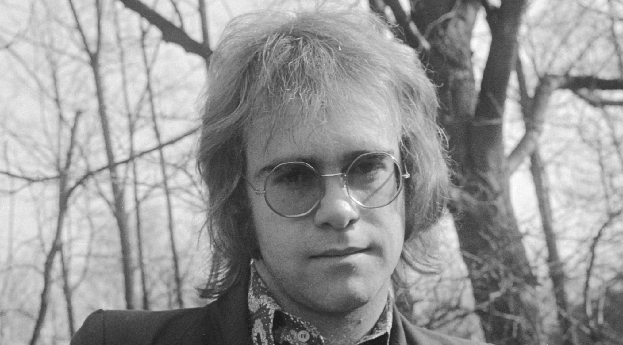 A black and white image of Elton John, one of the famous people before the fame, wearing round glasses.