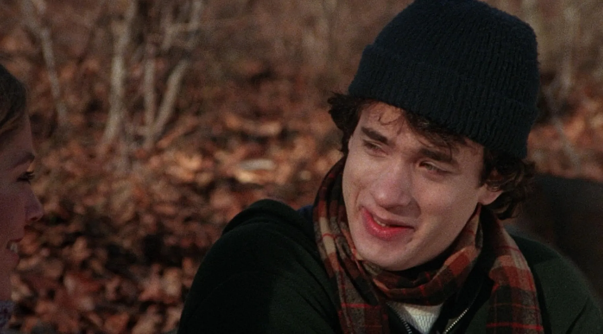 A young Tom Hanks starting his successful career, smiling subtly while wearing a beanie and a plaid scarf outdoors.