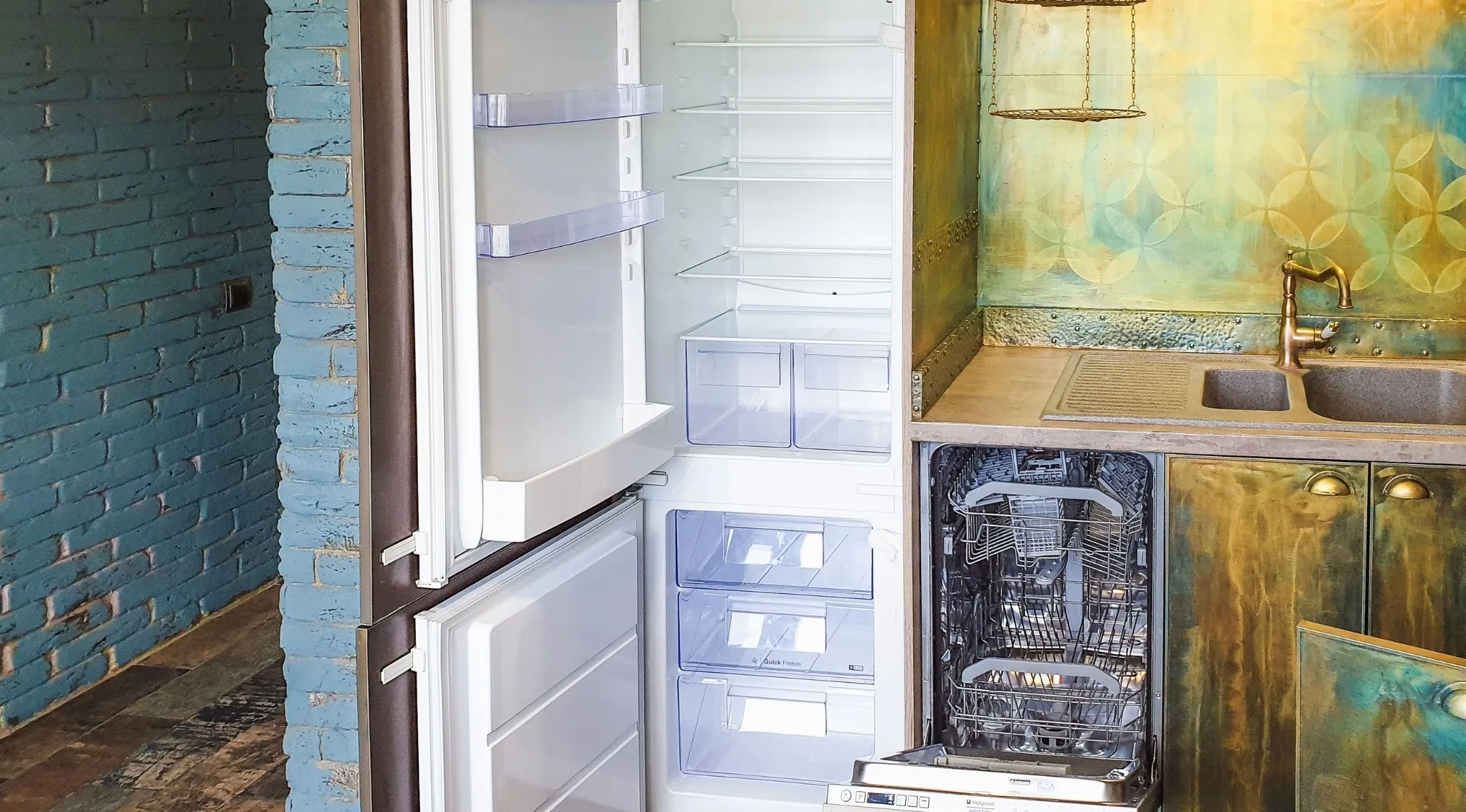 An open, empty refrigerator and dishwasher in a kitchen with blue brick and metallic green walls. Home economics of refrigerator temperature.