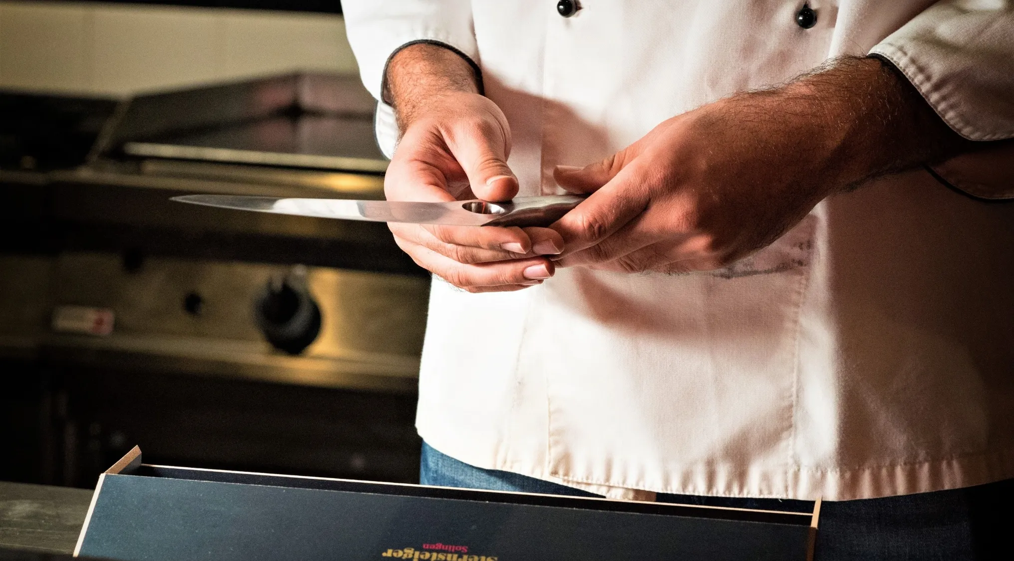 A chef in a white jacket carefully examines a chef's knife in his hands, preparing to demonstrate proper holding technique.