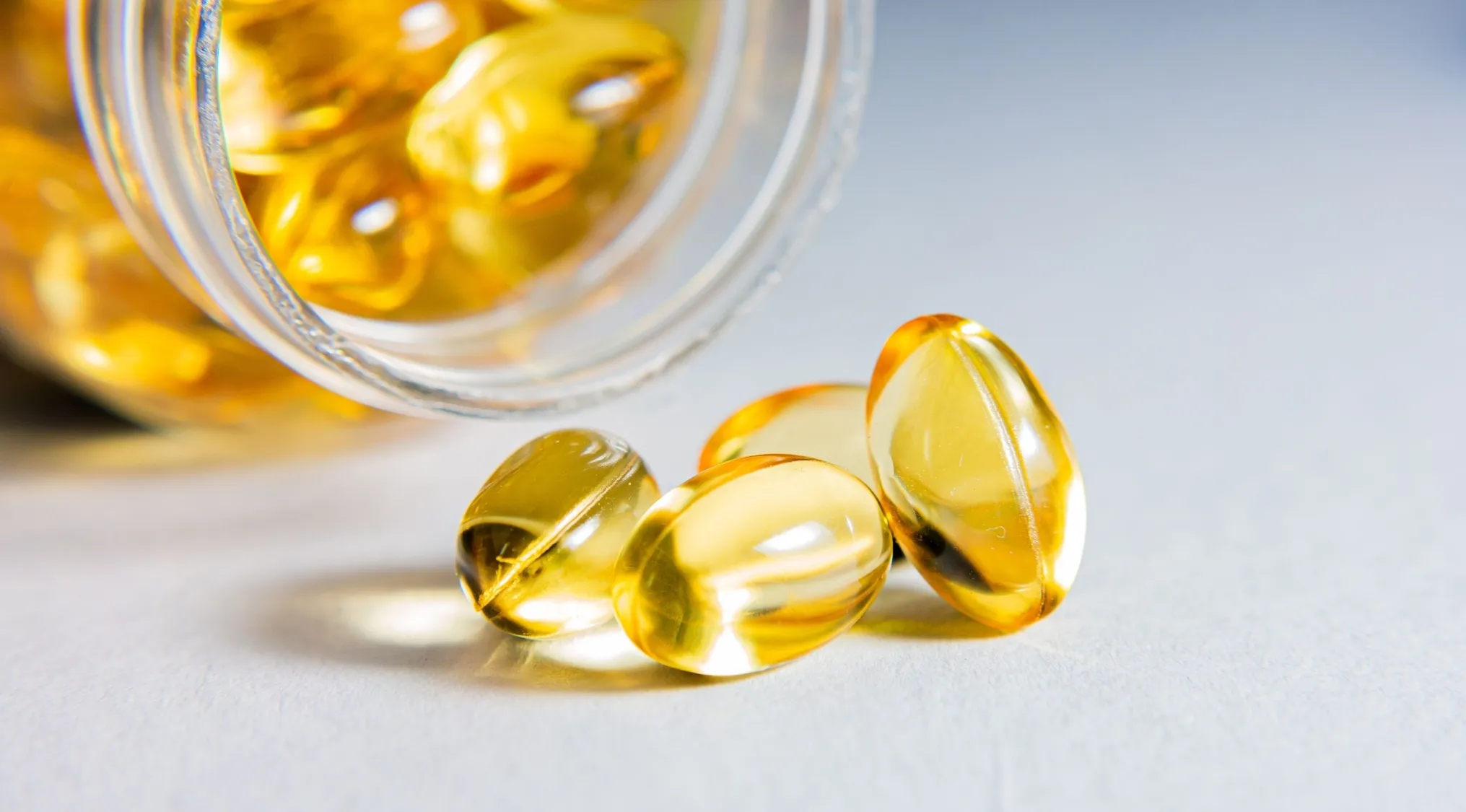 Bright yellow softgel Vitamin A capsules spilling from a glass jar onto a light surface.