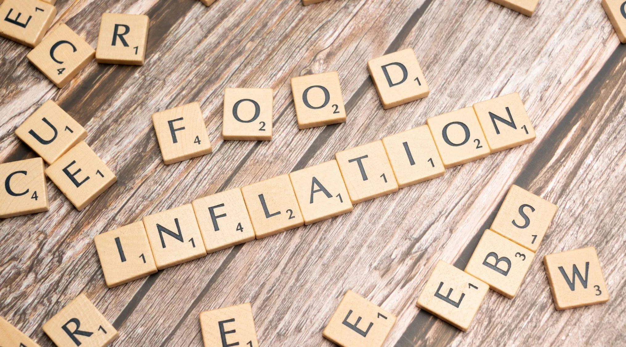 Scrabble tiles on wood grain spelling out FOOD INFLATION, representing economic principle of supply and demand.