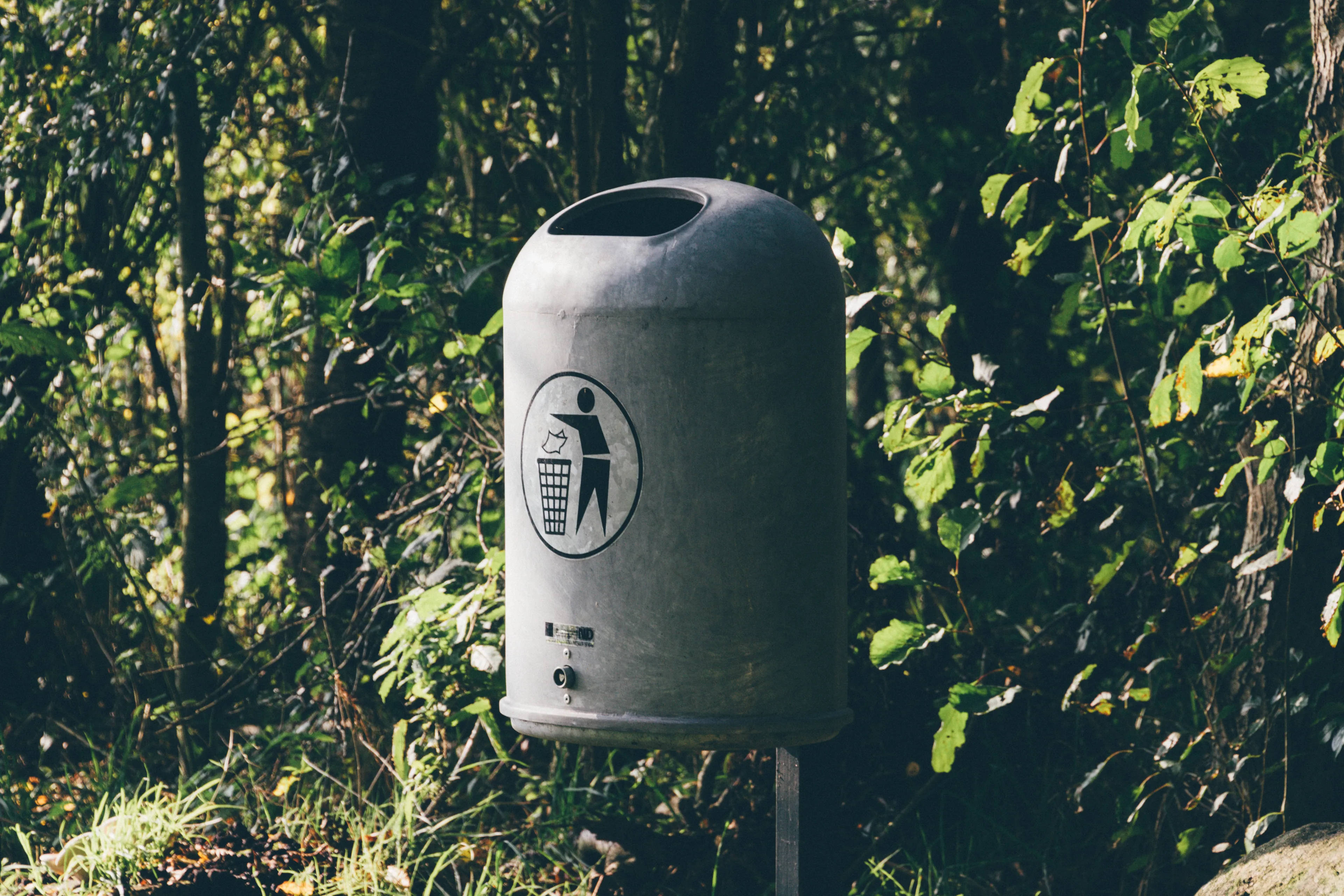 A grey trash bin, where Brits throw things away, in a park setting for American vs English Words Quiz.