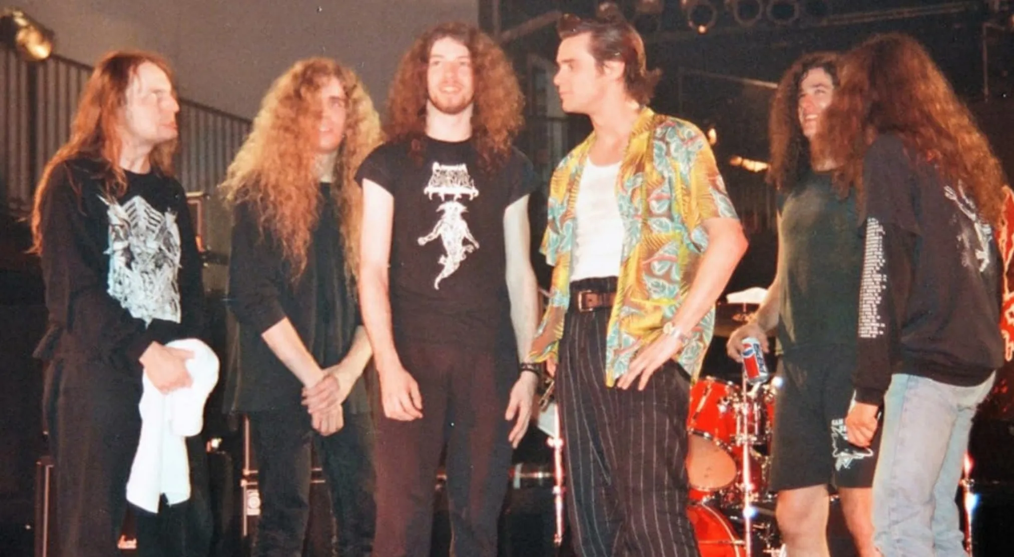 A behind the scenes image showing a group of men with long hair and Jim Carrey on stage. Guess the movie.