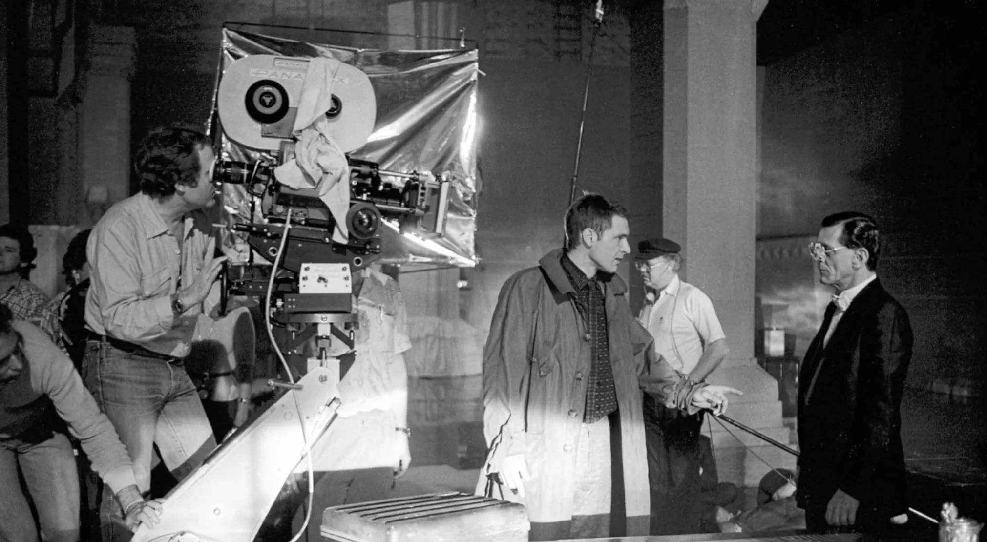 Black and white BTS image of a movie set with a cameraman, Harrison Ford in a trench coat, and an older man in a suit. Guess the movie.