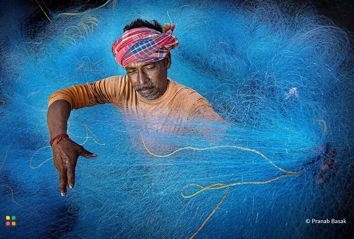 A man with a red and white turban sits surrounded by a vibrant blue fishing net, showcasing award-worthy photos.