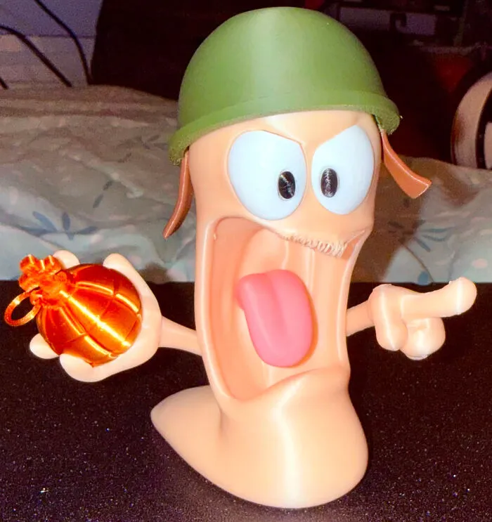 A 3D printed figurine of a cartoon worm in a military helmet, holding a glowing bomb and pointing, with an open mouth and tongue out.
