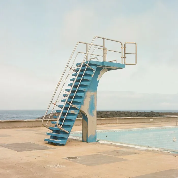 An empty blue diving board with stairs overlooking an abandoned swimming pool and the ocean. reFocus Contest photo capturing the world.