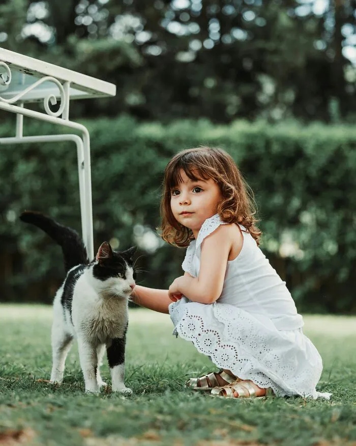 A young girl in a white dress squatting to pet a black and white cat outdoors, showing kids interacting with the world.